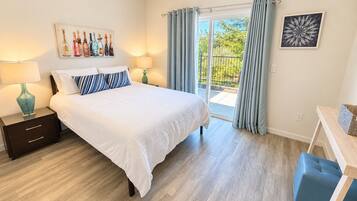 Un dormitorio con cama, mesitas de noche, una ventana con cortinas y vistas al exterior con vegetación, además de un cuadro en la pared.