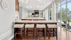 A modern kitchen with a central island, bar stools, pendant lighting, and large windows offering a view of the outdoors.