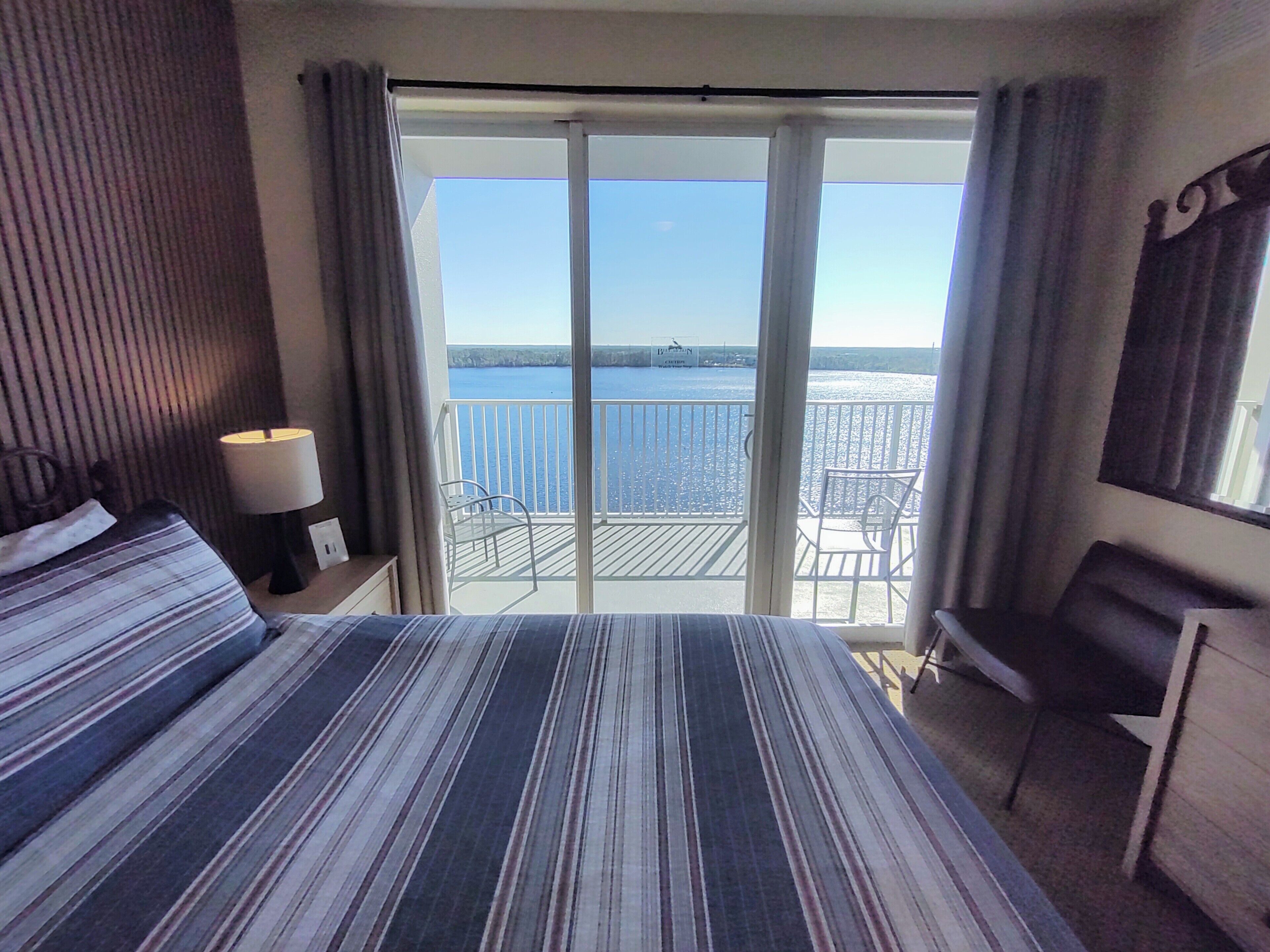 A hotel room with a large bed, a desk, a chair, and a balcony with a view of the sea.