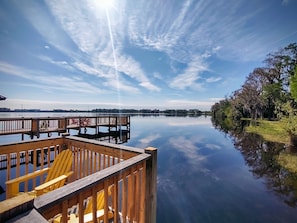 A calm lake with a wooden dock and chairs, surrounded by trees and buildings in the distance.