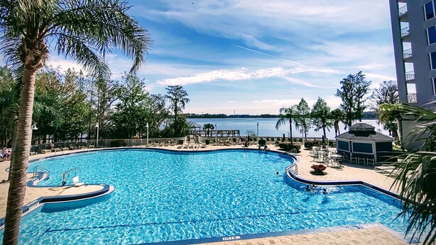 A swimming pool with a view of a body of water, surrounded by palm trees and a building. - DISNEY area CONDO 2 KING BEDS 2 BUNKS 2 BATHS Large 1350 sq ft Lake View 10th fl (Orlando)