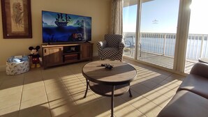 A living room with a TV, a Mickey Mouse plush toy, a patterned armchair, a round table, and a balcony with a view of the sea. - DISNEY area CONDO 2 KING BEDS 2 BUNKS 2 BATHS Large 1350 sq ft Lake View 10th fl (Orlando)
