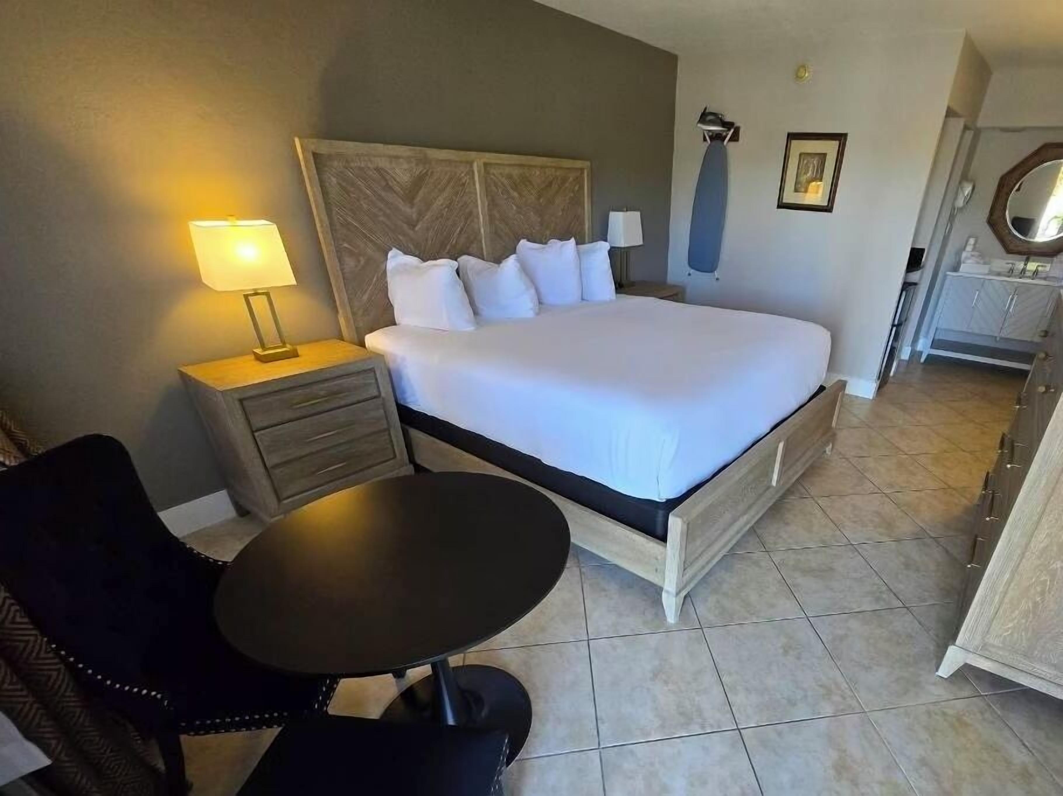 A hotel room with a bed, a desk, a chair, a lamp, a mirror, and a dresser.