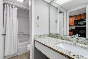 Ein Badezimmer mit einer weißen Badewanne, einem Waschbecken mit einer Granit-Arbeitsplatte und einem Spiegel, der in den Küchenbereich blickt.