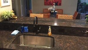A kitchen with a granite countertop, stainless steel sink, and a dining area with wicker chairs and a table. - RENOVATED PROPERTY!!  Book now! E101 (Waikoloa)