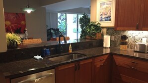 A modern kitchen with a stainless steel dishwasher, wooden cabinets, a black countertop, a sink, and a hanging light fixture. - RENOVATED PROPERTY!!  Book now! E101 (Waikoloa)