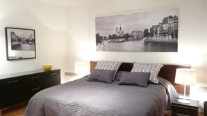 Un dormitorio con una cama grande, una mesita de noche con lámpara, un aparador y dos cuadros enmarcados en la pared.
