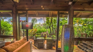 A covered outdoor seating area with wicker furniture, a table with a vase of flowers, and a view of lush greenery.