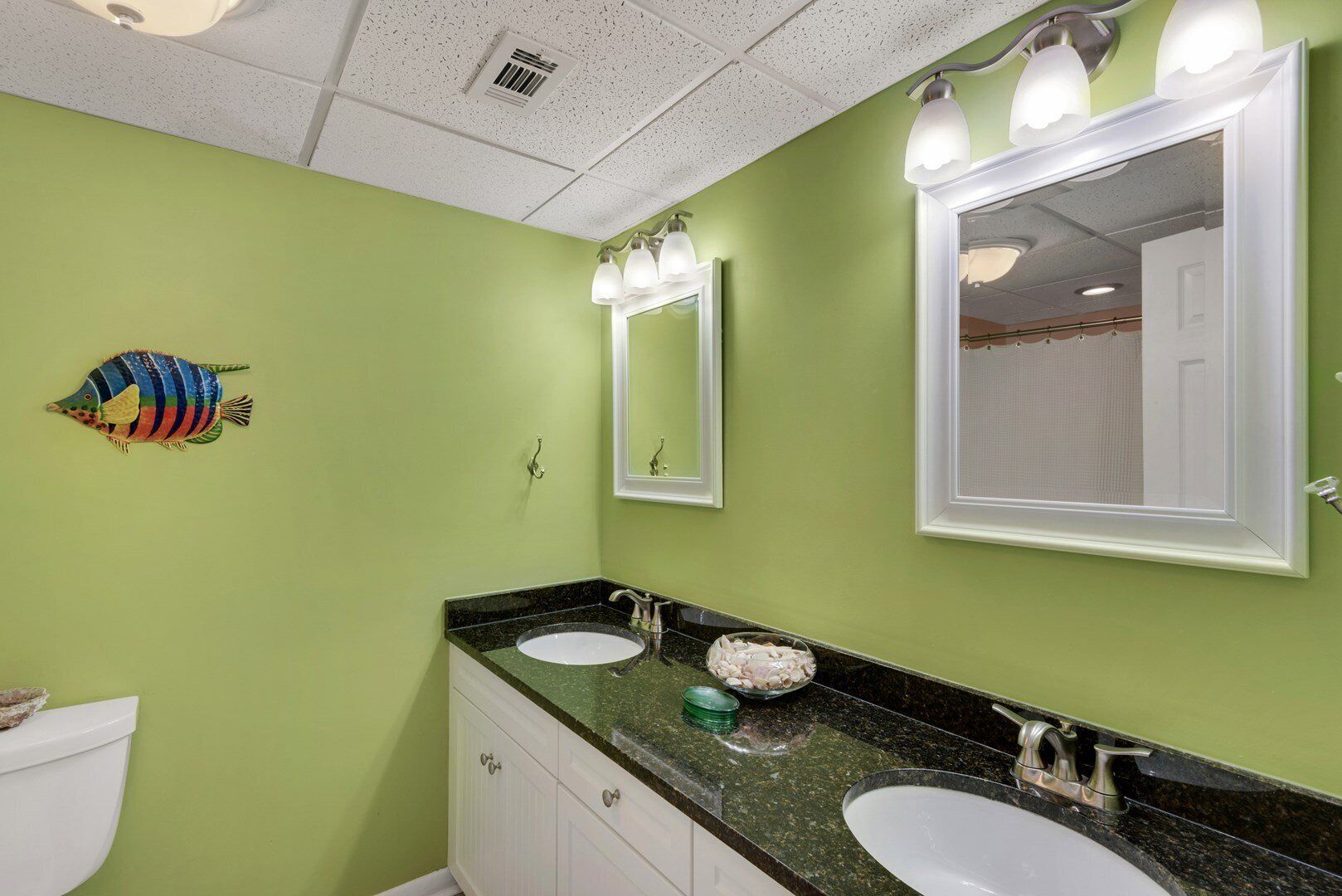 A bathroom with green walls, two sinks, a mirror, and a ceiling vent.