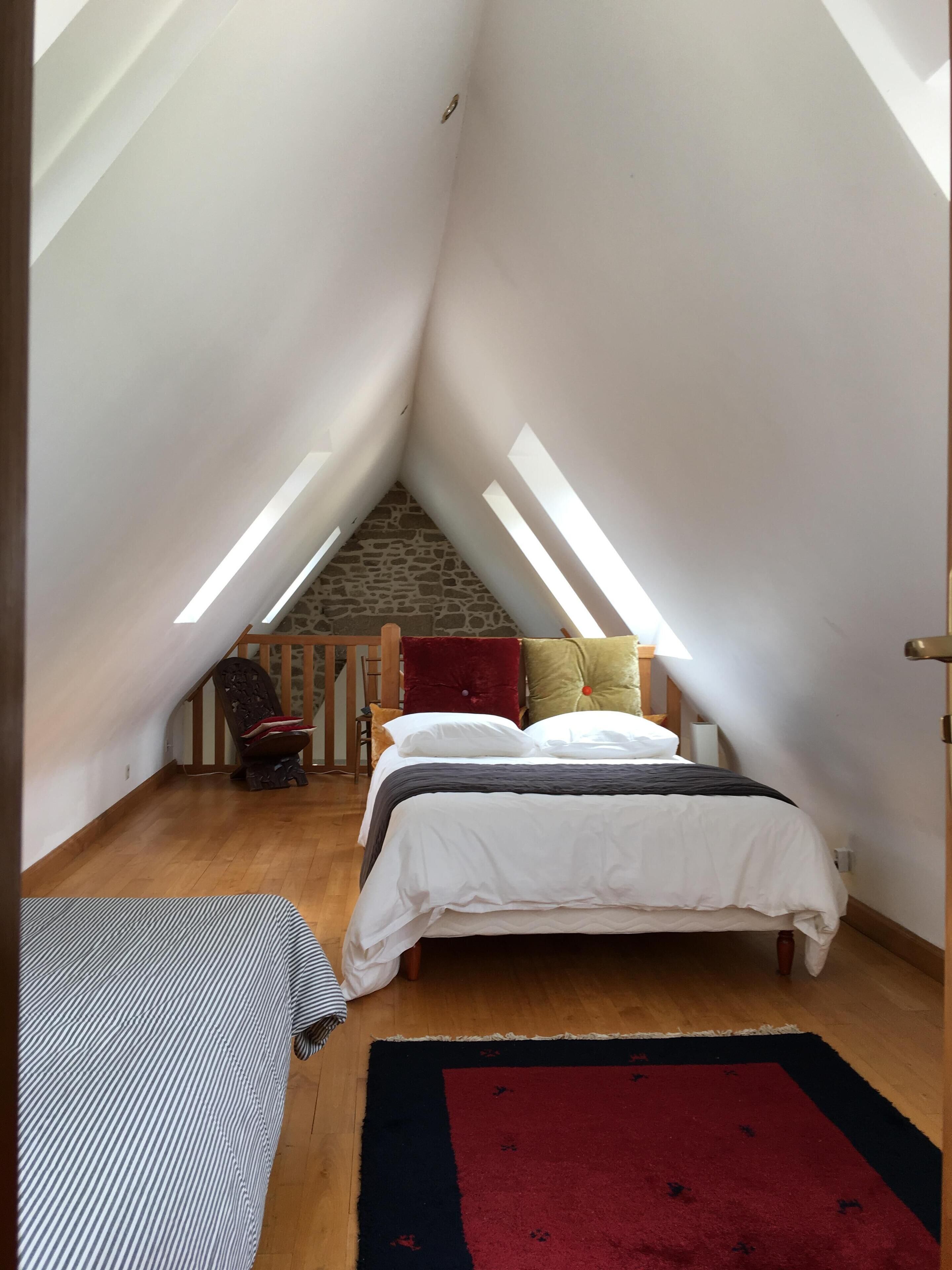 A bedroom with a sloped ceiling, a bed, a red and black rug, and a stone wall.