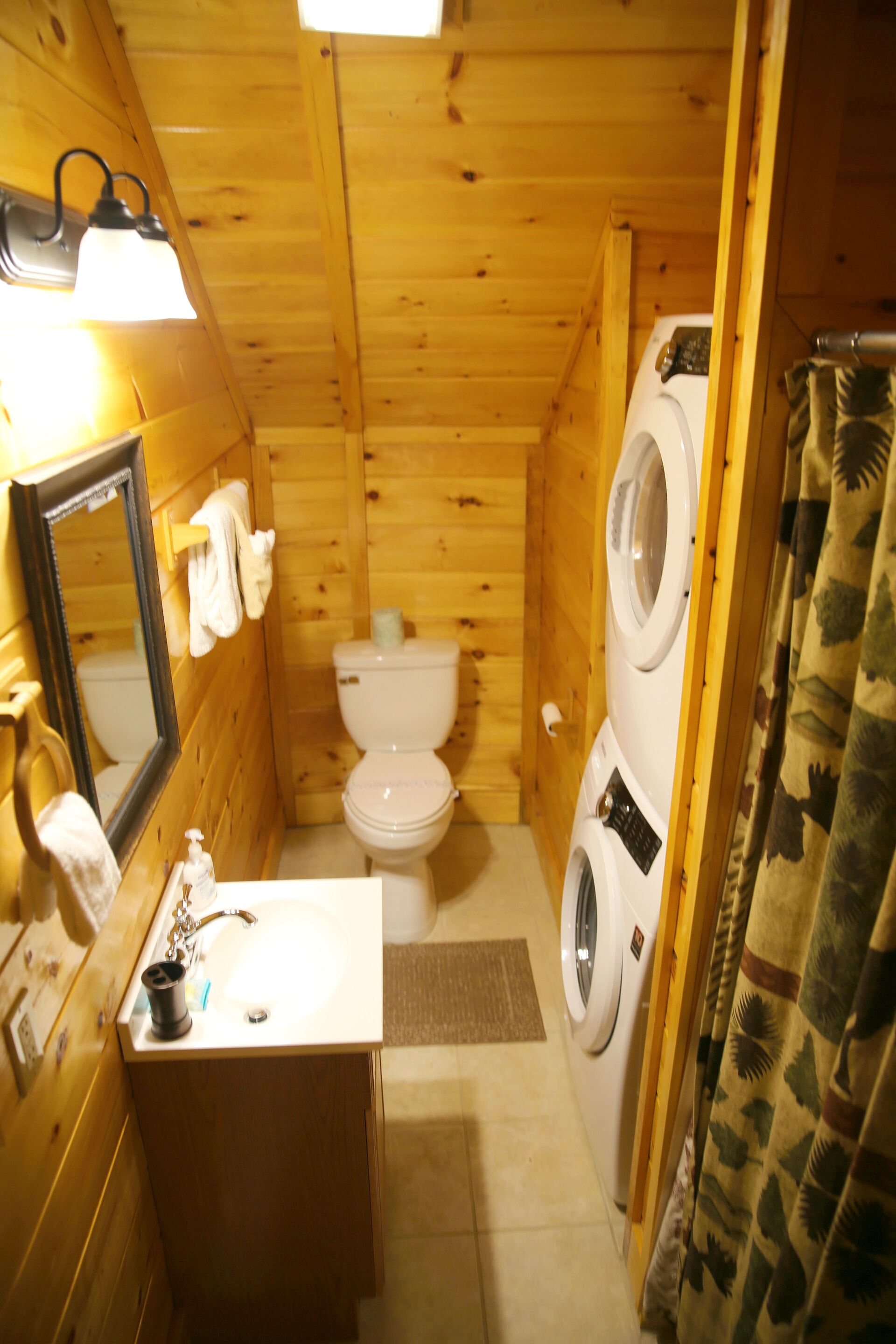 Een klein houten badkamertje met toilet, wasmachine, wastafel en spiegel.