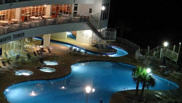 En nattbild av ett hotells poolområde med flera pooler, solstolar och en trappa som leder upp till en övre våning.