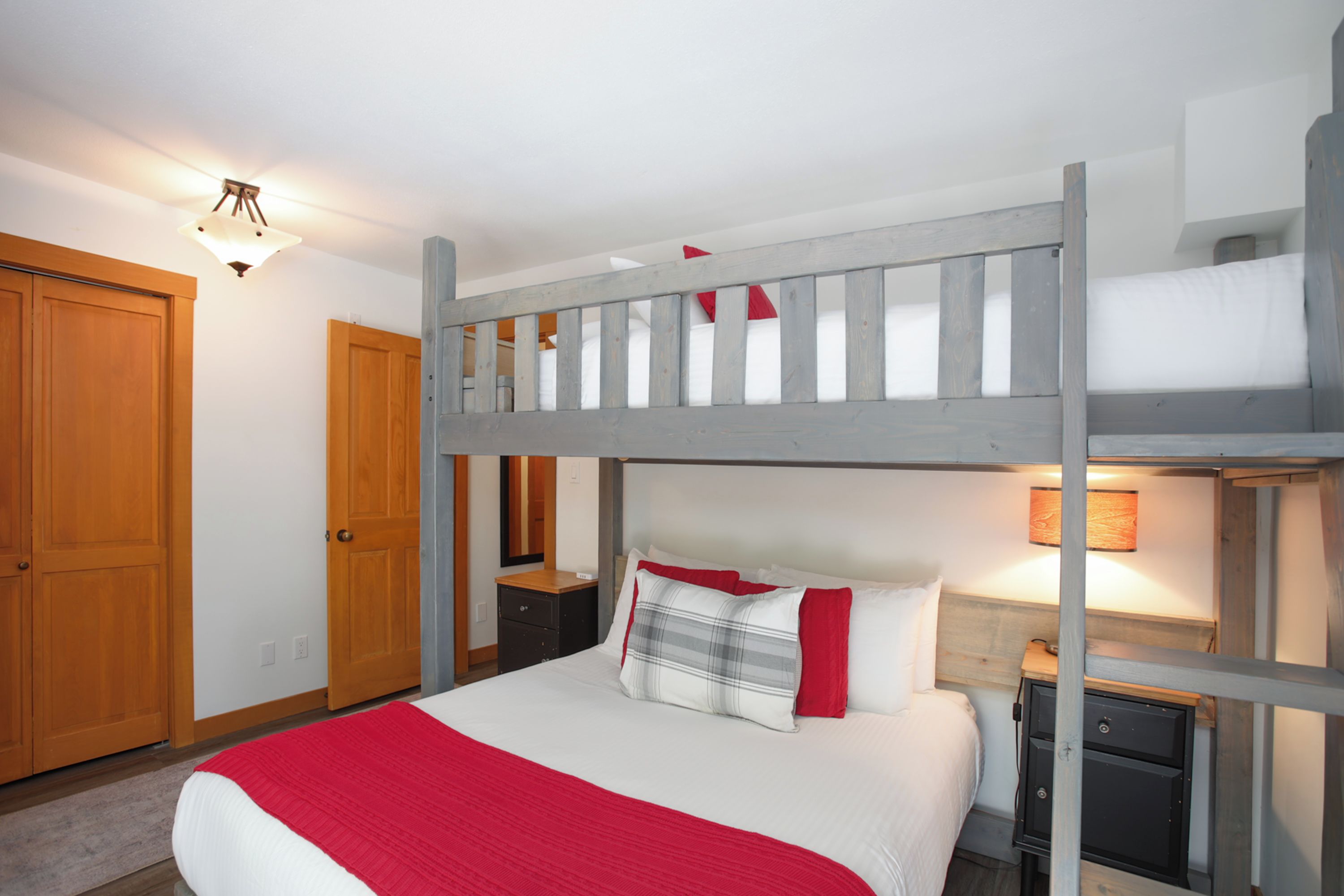 Una cama litera con una colcha roja, un armario de madera y una lámpara de noche.