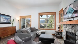 A modern living room with a grey sectional sofa, a fireplace, and large windows offering a view of the outdoors. - Mountain Star 16 - Ski in Walk out, Private Hot Tub, Fireplace (Whistler)