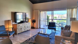Una sala de estar moderna con una televisión de pantalla plana, un ventilador de pie y un amplio ventanal con vistas a edificios y zonas verdes.