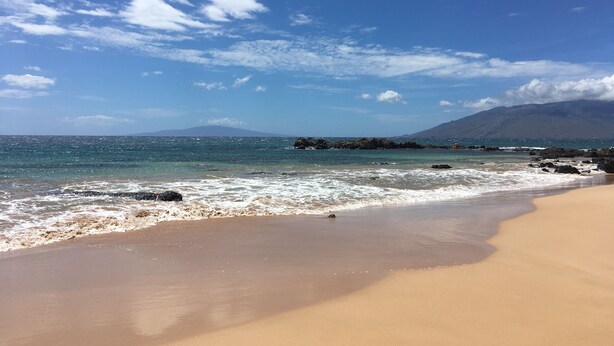 Una playa con cielos despejados, aguas tranquilas y una montaña distante.