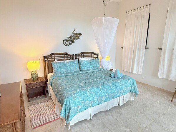 A bedroom with a bed, a nightstand, a lamp, a mosquito net, and a wall decoration. - Bask in Beauty: Anole Offers Unmatched North Shore Views and a Pool Perfect for Relaxation (Cinnamon Bay)