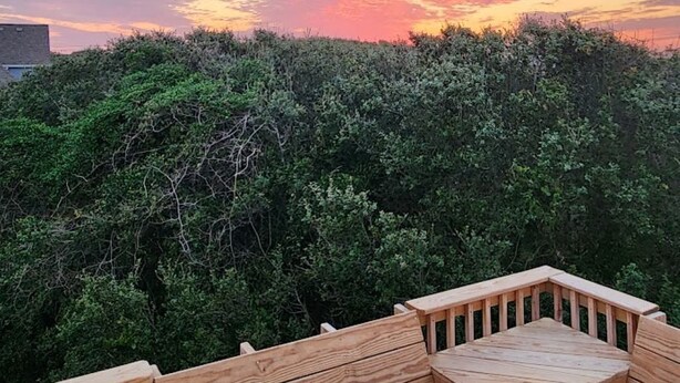 A wooden deck with a table and benches, surrounded by dense foliage and a sunset sky. - Cozy Cottage in Kitty Hawk with Gorgeous Ocean Views! (Kitty Hawk)