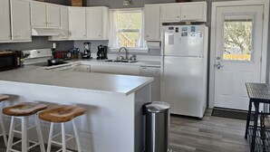 A modern kitchen with white cabinets, a central island with stools, a stainless steel trash can, and a refrigerator. - Cozy Cottage in Kitty Hawk with Gorgeous Ocean Views! (Kitty Hawk)