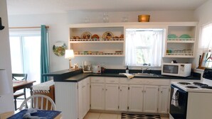 A compact kitchen with white cabinets, a microwave, a stove, and a sink. There is a dining table with chairs and a blue mat on it. - High Hedges Cottage  - Close to downtown Mt Dora in quiet neighborhood (Mt Dora)