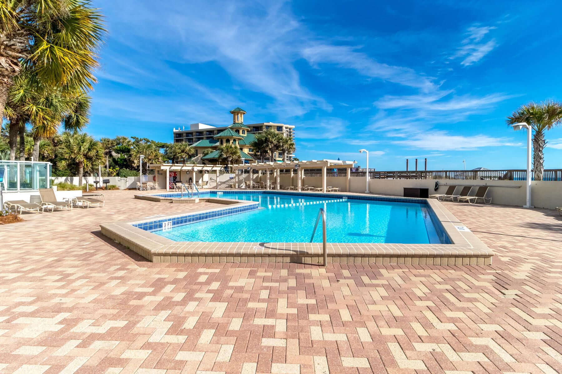 A rectangular swimming pool with a tiled deck, surrounded by lounge chairs and palm trees.