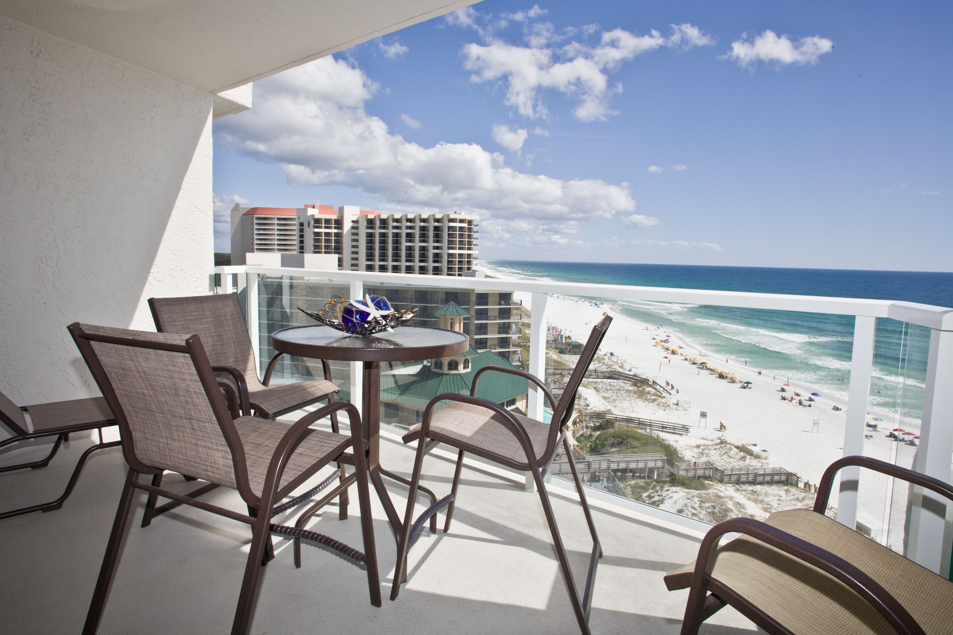 A balcony with wicker chairs and a table, overlooking a beach with people and buildings in the background.