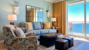 A living room with a blue sofa, patterned armchair, and ottomans, a large window with curtains, and a view of the ocean. -  Boardwalk Beach Resort, Ocean/Beach front, Loaded w Amenities, Park your car!! (Panama City Beach)