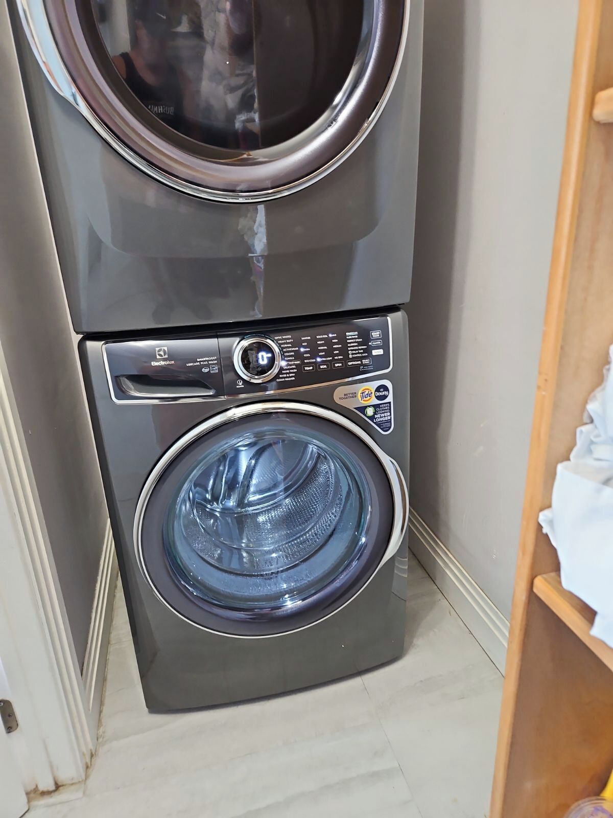 A stacked washer and dryer unit with a control panel, located in a corner with a wooden shelf nearby.