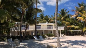 Une propriété située en bord de mer, comprenant un bâtiment à deux étages, un balcon et une plage de sable avec des chaises longues.