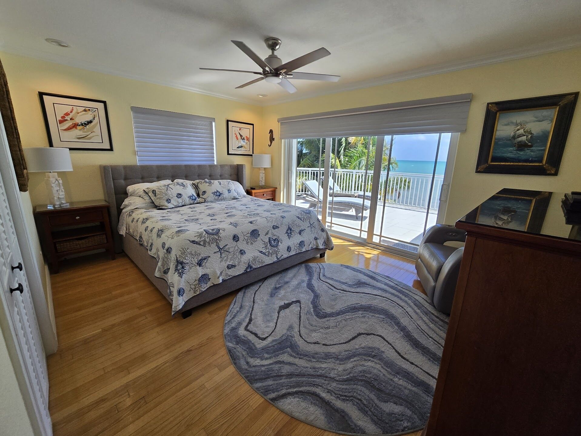 A bedroom with a bed, a ceiling fan, a nightstand, a lamp, a window with blinds, and a view of the ocean through a balcony.