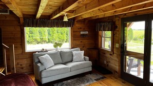 A cozy cabin interior with a grey sofa, wooden walls, a 'Welcome' sign, and a view of greenery outside. - Heritage hideaway-Secluded Log Cabin (Chatfield)