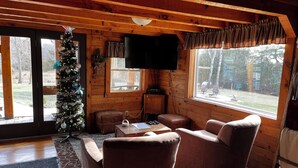 A cozy cabin interior with a wooden interior, a Christmas tree, leather chairs, a flat-screen TV, and a large window offering a view of the outdoors. - Heritage hideaway-Secluded Log Cabin (Chatfield)
