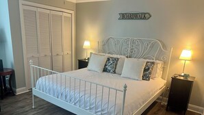 Un dormitorio con estructura de cama metálica blanca, suelo de madera, mesita de noche con lámpara, una mesilla auxiliar y un armario con puertas persianas.