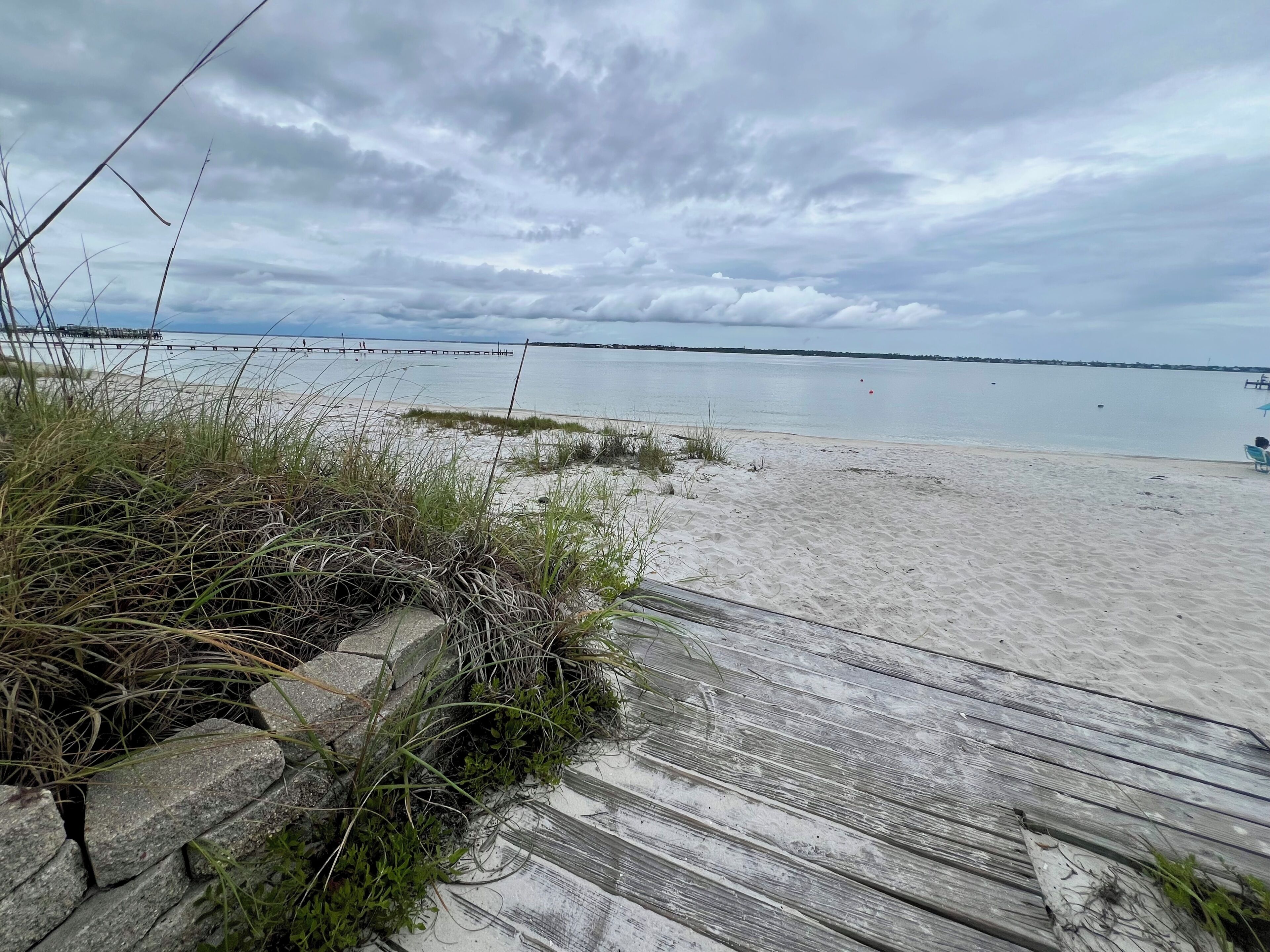 A beach with a wooden boardwalk, grassy areas, and a calm sea under a cloudy sky.