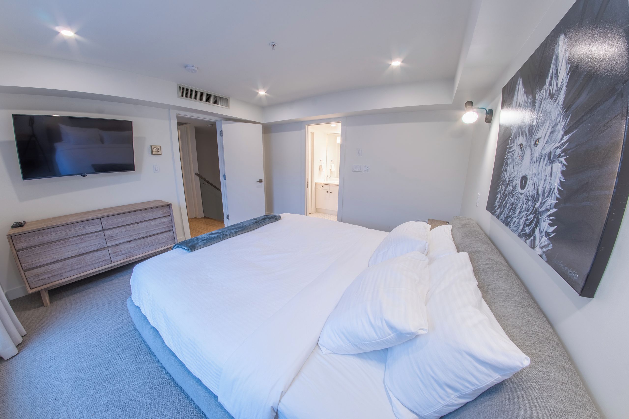 A bedroom with a large bed, a TV mounted on the wall, a dresser, and a painting.