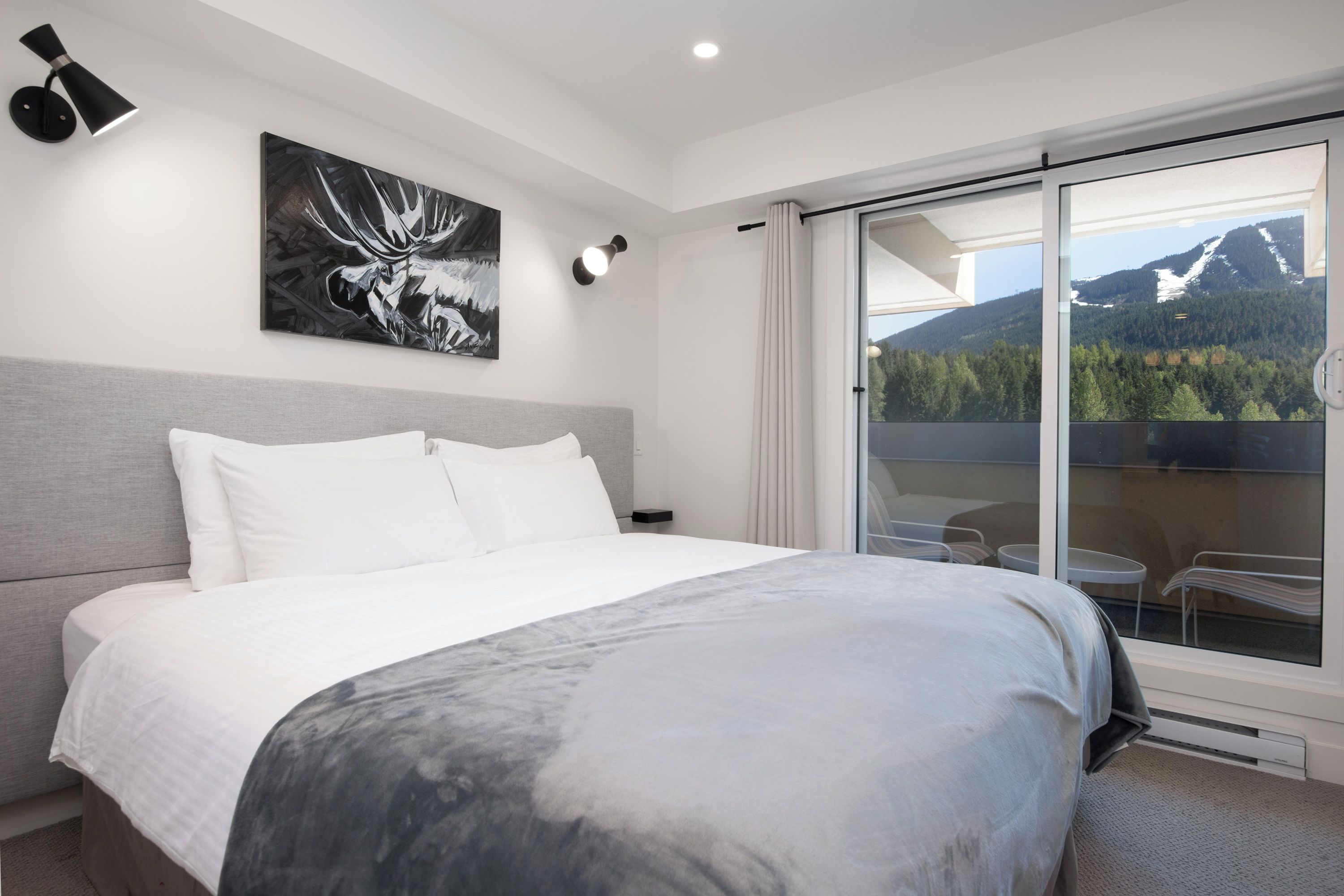 A modern hotel room with a large bed, a painting of a moose, a balcony with a mountain view, and a sliding glass door.