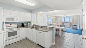 A modern kitchen with white cabinetry, a central island, and a dining area with a view of the ocean. - Nautical Oceanfront Condo, Jacuzzi Tub, Indoor Pool | Crescent Shores - S 1711 (North Myrtle Beach)