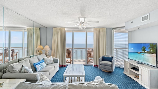 A spacious living room with a sofa, armchairs, a coffee table, and a TV. The room has large windows with curtains, a ceiling fan, and a view of the ocean. - Nautical Oceanfront Condo, Jacuzzi Tub, Indoor Pool | Crescent Shores - S 1711 (North Myrtle Beach)