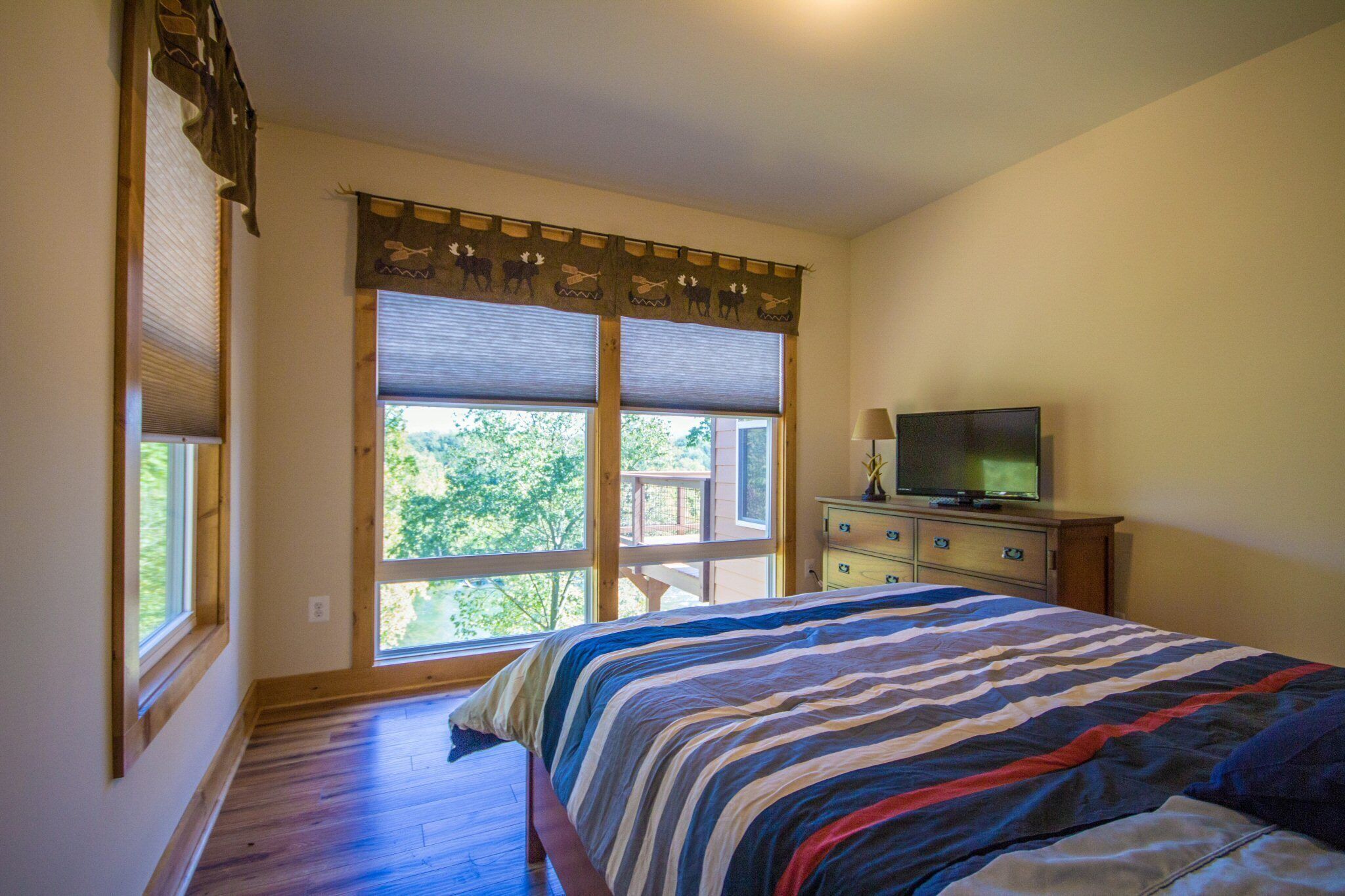 A bedroom with a large bed, a dresser, a television, and a window with a view of trees.