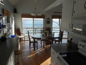 A kitchen with a large window overlooking the ocean, wooden flooring, and a dining area with chairs and a table. - Lakefront property close to town. Spectacular views and location. (Skaneateles)