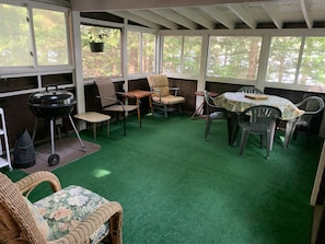 A covered patio area with green artificial turf, a barbecue grill, wicker furniture, plastic chairs, and a table with a floral tablecloth.