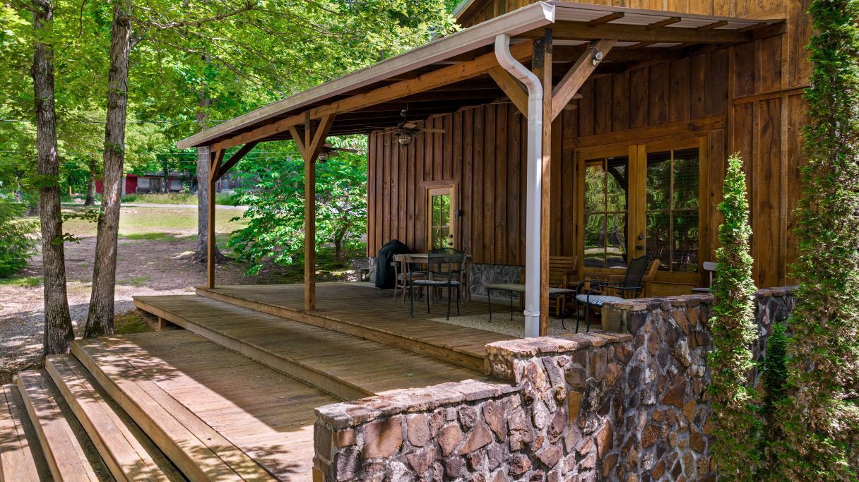 A rustic wooden cabin with a porch, stone walls, and a table and chairs set up for outdoor dining.