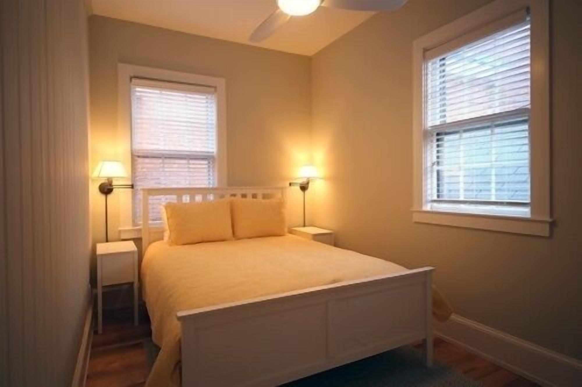 A bedroom with a bed, two bedside lamps, and two windows with blinds.