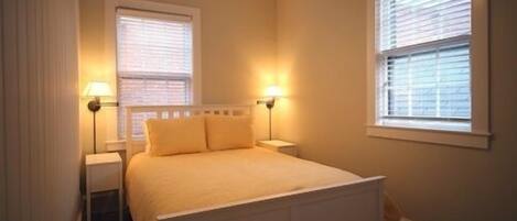 A bedroom with a bed, two bedside lamps, and two windows with blinds.