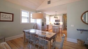 A dining area with a wooden table and chairs, a kitchen in the background, and a framed picture on the wall.