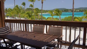 Eine Holzterrasse mit Tisch und Stühlen mit Blick auf einen Strand, an dem Palmen stehen und türkisfarbenes Wasser zu sehen ist.