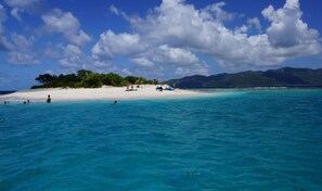 Una playa con aguas turquesas cristalinas, una orilla de arena blanca y una pequeña isla con árboles y algunas personas.
