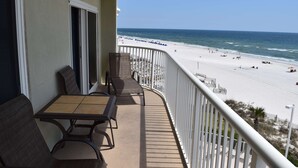 A balcony with a table and chairs, overlooking a beach with people and umbrellas. - Beautiful Ocean Front Condo in Orange Beach....Affordable Prices (Orange Beach)