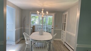 Un coin repas avec une table ronde, des chaises blanches, un lustre et une vue sur l’extérieur, visible à travers une porte vitrée.