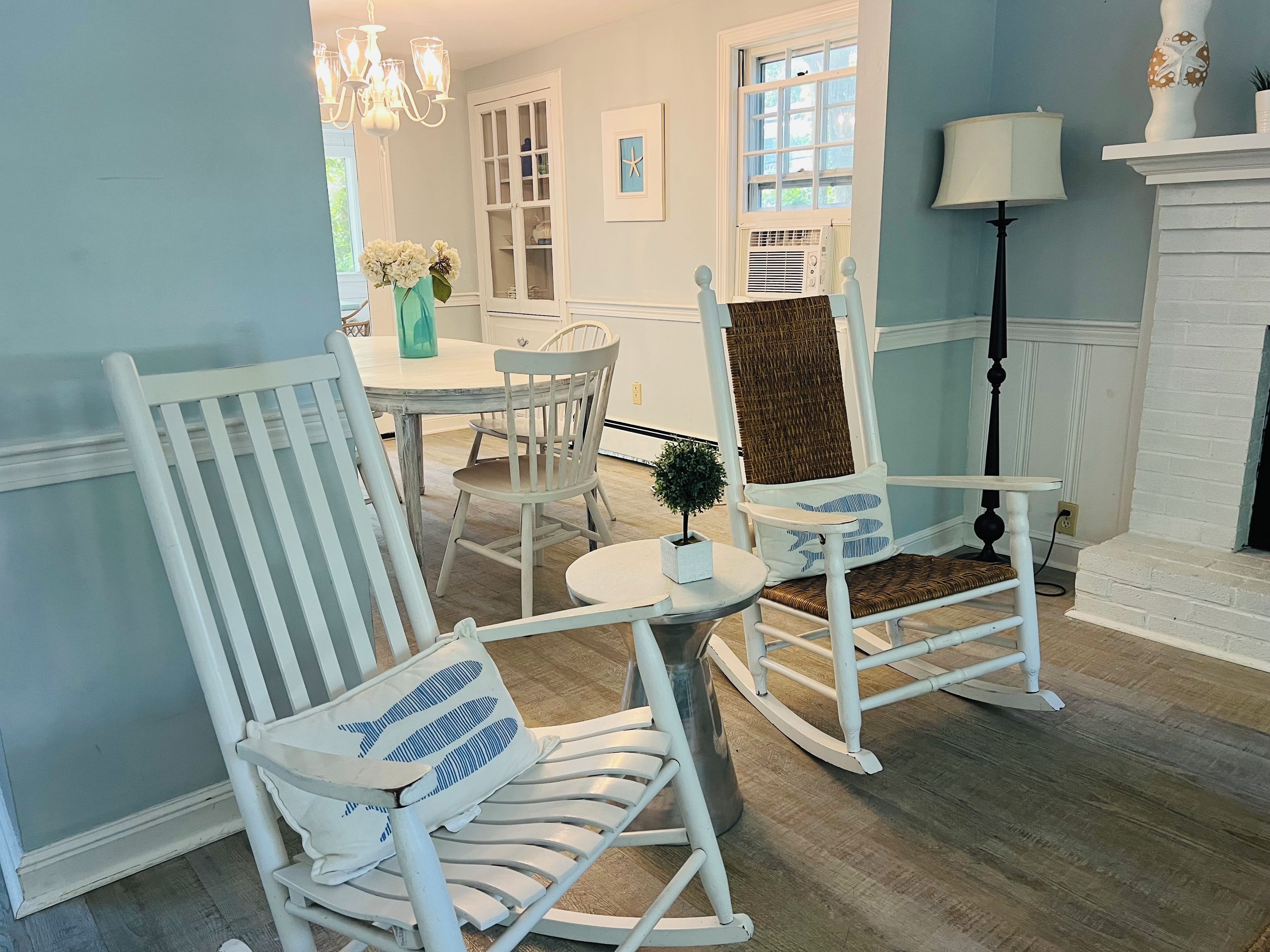 A room with a white rocking chair, a small table with a plant, and a dining area in the background.